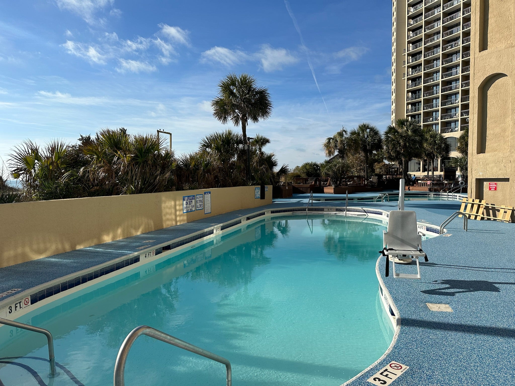 Myrtle Beach Cove - After - with rubber surfacing - Pool area with lounge chairs, palm trees, and a high-rise building in the background.