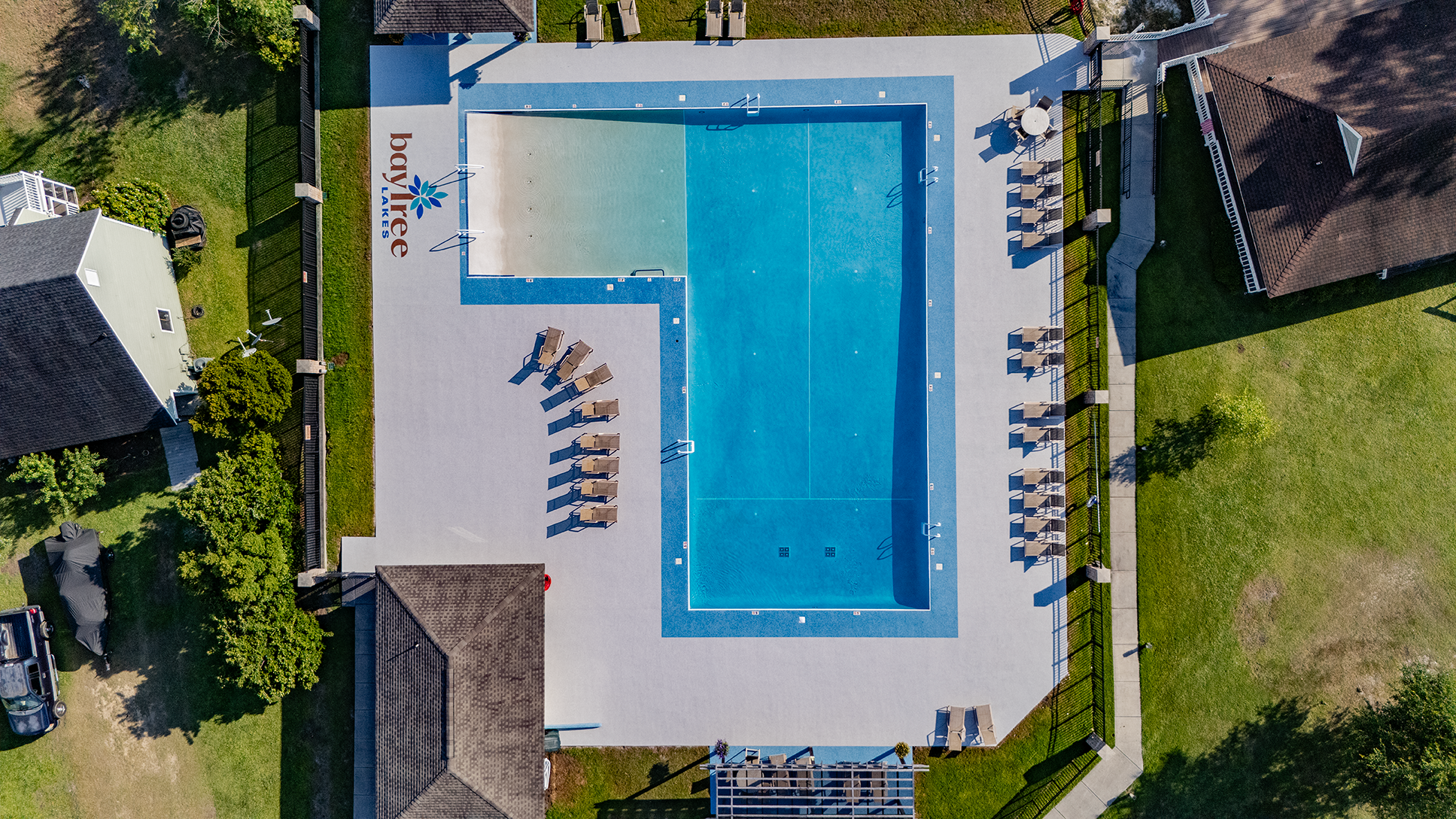 Aerial view of a pool with lounge chairs and umbrellas surrounded by grass and houses.