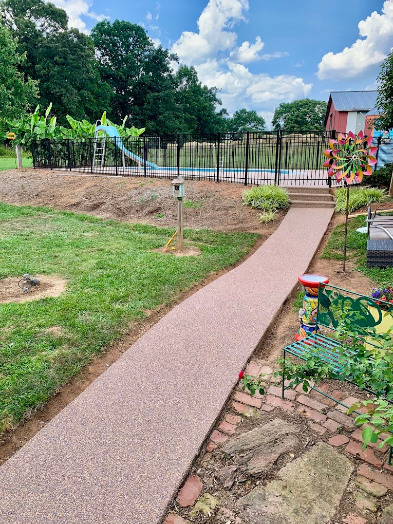 Backyard with a pathway leading to a pool area with a slide, surrounded by trees and a clear sky. Rubber surfacing is installed in the walkway.