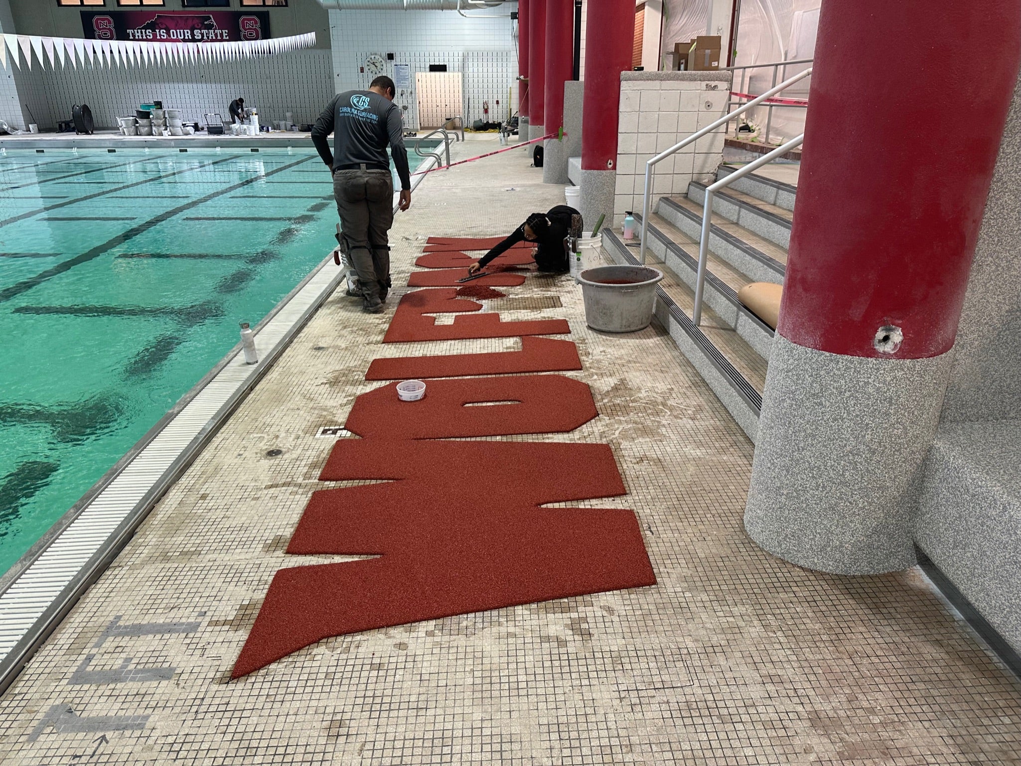 NC State Aquatic Center - During Rubber Surfacing Installation - Workers installing red letters by an indoor pool.