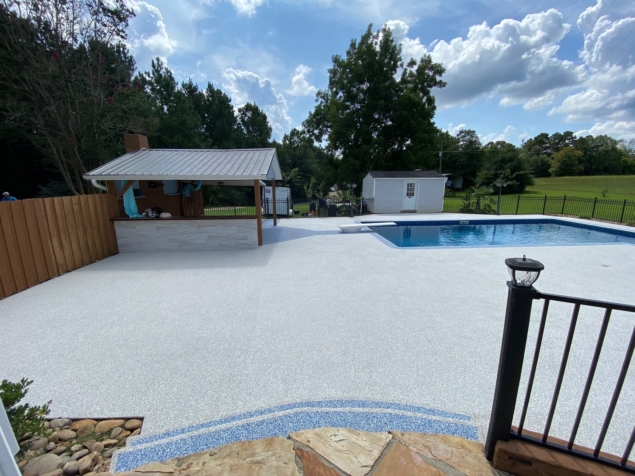 Residential - Backyard with pool, deck, and surrounding trees under a blue sky. After image - White / Cream Rubber Surfacing installed.