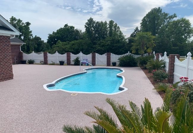 Swimming pool in a backyard with trees and a white fence. Rubber surfacing installed