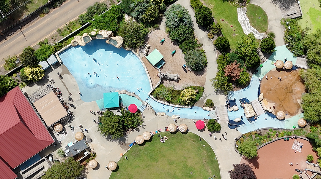 Aerial view of a large outdoor pool area with various pools and landscaping. Rubber surfacing around pool area.
